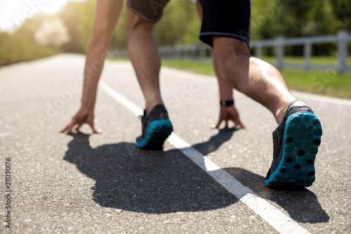 Close-up of male feet in crouch start on asphalt track outside and turning back to camera. He is bending body and putting hands on road while feeling concentrated. Guy is wearing trainers and watch