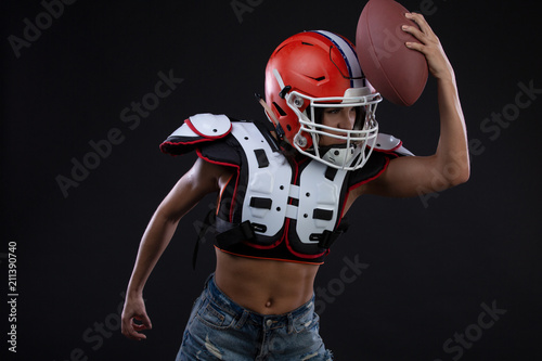 Sportive serious woman in helmet of rugby player holding ball in stuio on black background.