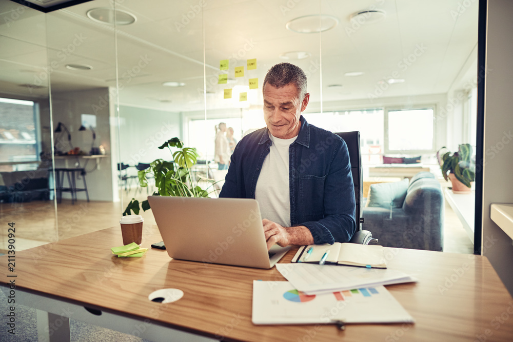 Smiling mature businessman sitting at his desk using a laptop Smiling mature businessman sitting at his desk using a laptop