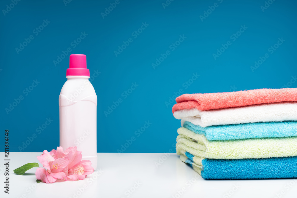 Close up of pink bottle of laundry liquid with beautiful flowers aside. Clean washed towels lying in right side on white table Close up of pink bottle of laundry liquid with beautiful flowers aside. Clean washed towels lying in right side on white table