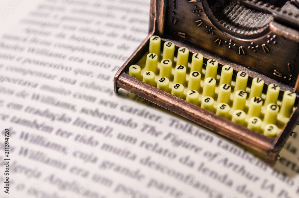 Small typewriter with keyboard in focus over a page of a book. Decoration  item. Concepts of reading, writing, old times, past. Close up. Blurred  background. Stock Photo