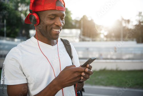 Positive male athlete listening music while typing in mobile. He standing outdoor. Copy space