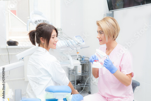A female dentist explains to a female patient the process of brushing teeth on a mock-up. Both look at the camera