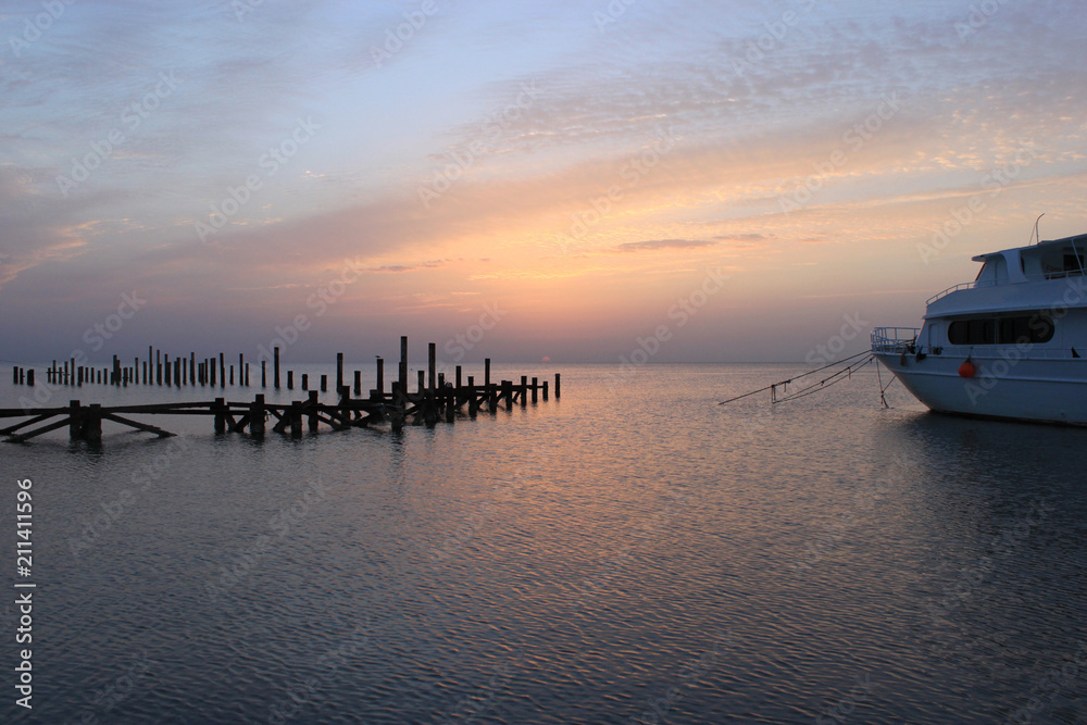 Sunrise on the sea. Sky is painted in shades of pink. Wooden piles of old destroyed pier sticking out of water and ship. Early in the morning sun appears over the horizon.