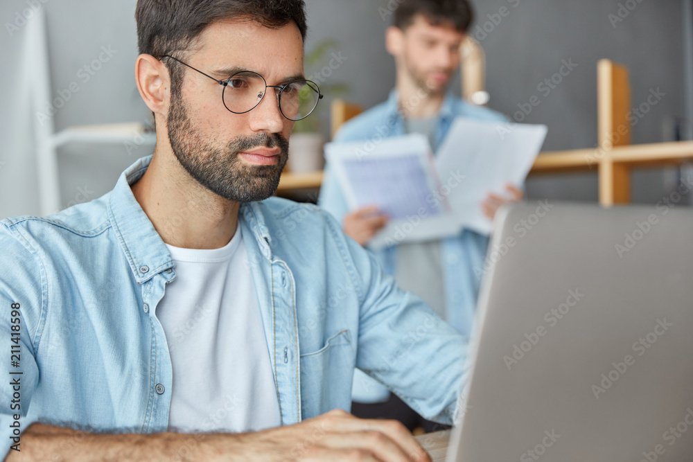Obraz premium Zdjęcie dwóch programistów instalujących nową aplikację, współpracujących w nowoczesnym biurze. Odnoszący sukcesy biznesmen o atrakcyjnym wyglądzie, pracuje na laptopie, skupia się na informacjach na stronie internetowej, korzysta z internetu