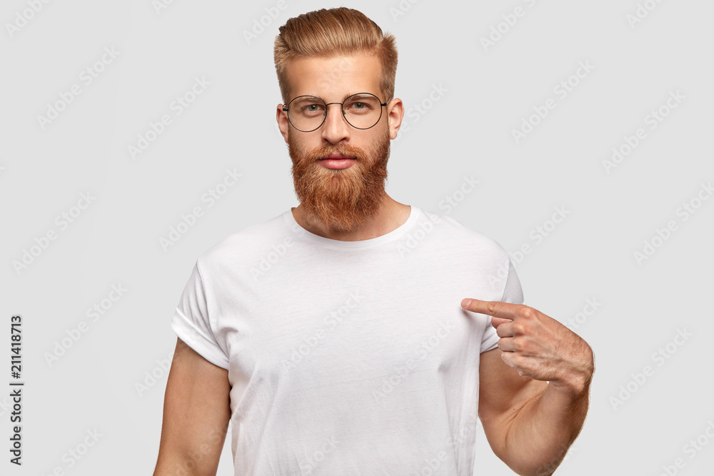 People, advertisement and clothing concept. Serious male hipster with trendy haircut and red beard, indicates at blank space of his t shirt, shows place for your logo, stands indoor, wears spectacles People, advertisement and clothing concept. Serious male hipster with trendy haircut and red beard, indicates at blank space of his t shirt, shows place for your logo, stands indoor, wears spectacles