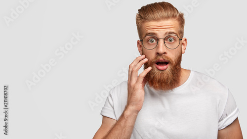 Astonished attractive male has ginger long beard, wonders sudden news, keeps mouth slightly opened, stares at camera, wears casual clothes and spectacles, poses against white wall with blank space