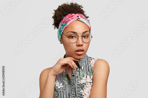 Indoor shot of pretty female with Afro hairstyle  looks seriously and mysterioulsy at camera  feels confident or self assured  demonstrates dark healthy skin  wears round glasses and headband