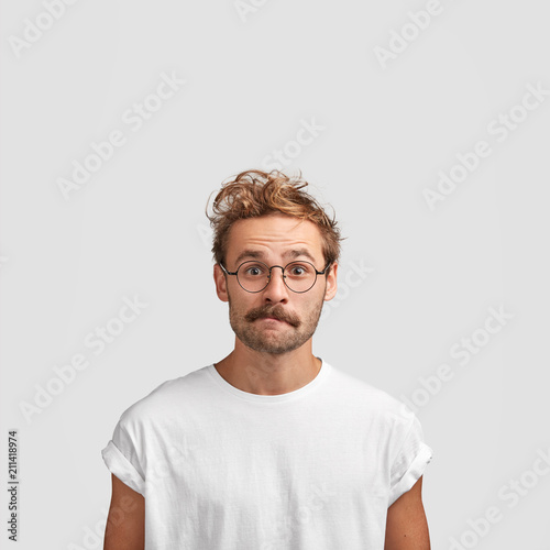 Vertical shot of puzzled European male with curly haircut, purses lips and looks in bewilderment, surprised to see big sales, dressed in casual white t shirt, isolated over studio background