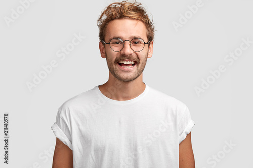 Horizontal shot of happy hipster male with toothy smile, wears casual white t shirt and glasses, being in good mood after unforgettable journey with girlfriend, isolated on white background. photo
