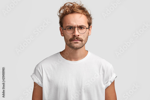 Indoor shot of serious man with grumpy expression, being discontent with noisy neighbours, dressed in casual white t shirt and spectacles, poses indoor. Attractive hipster has strict look and mustache