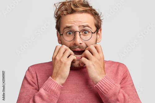 Emotive depressed young embarrassed man bites fingernails in panic, looks with nervous expression, going to cry with worried look, recieves negative awful news, poses against white background.