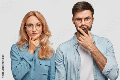 Horizontal shot of thoughtful male and female colleagues hold chins and being concentrated on solving problem, look directly into camera. Blonde middle aged female and her brother work as team