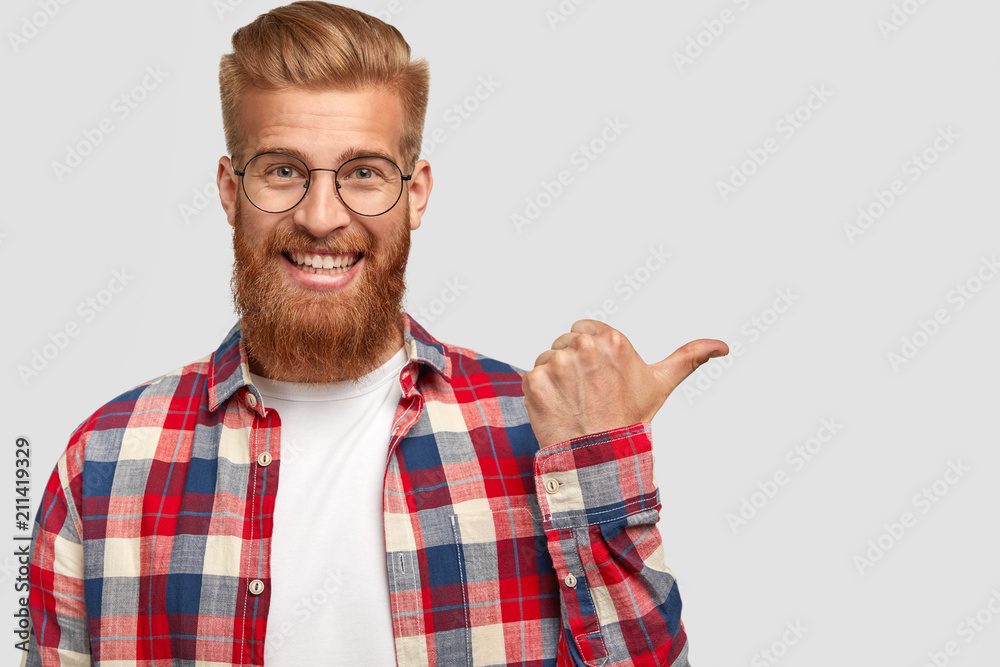 Naklejka premium Happy positive male with long thick ginger beard and mustache, has friendly smile, dressed in checkered shirt, happy to advertise something on blank space. People, style and promotion concept
