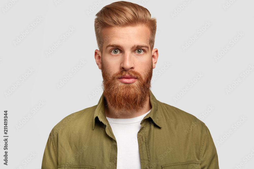Studio shot of handsome hipster has ginger beard, trendy hairstyle, looks seriously directly at camera, contemplates about future job, dressed in stylish shirt, poses against white studio wall Studio shot of handsome hipster has ginger beard, trendy hairstyle, looks seriously directly at camera, contemplates about future job, dressed in stylish shirt, poses against white studio wall