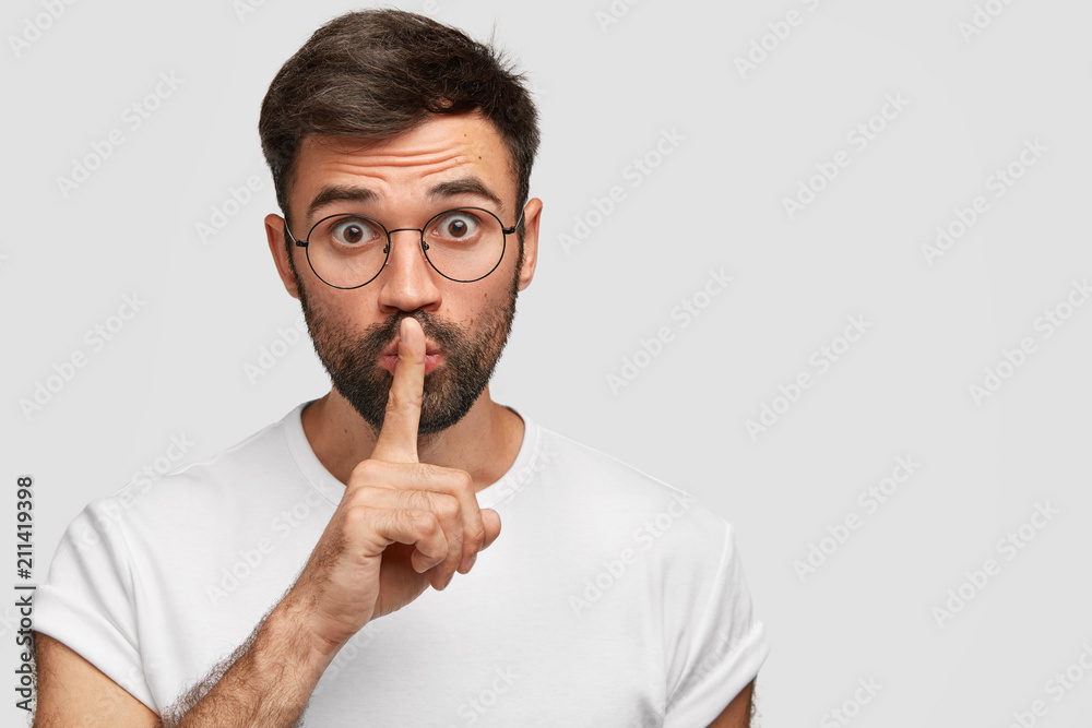 Studio shot of surprised young Caucasian male with thick beard, makes silence gesture, looks with bewilderment as afraids somebody knows his secret, dressed casually, isolated on white wall. Studio shot of surprised young Caucasian male with thick beard, makes silence gesture, looks with bewilderment as afraids somebody knows his secret, dressed casually, isolated on white wall.