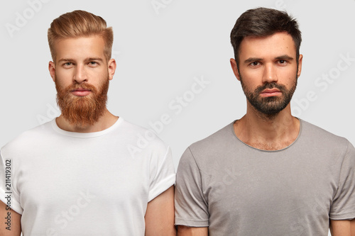 Bearded serious male friends with trendy haircut, stand close to each other, think where spend free time, dressed casually. Two stylish young men colleagues collaborate for creating common project photo