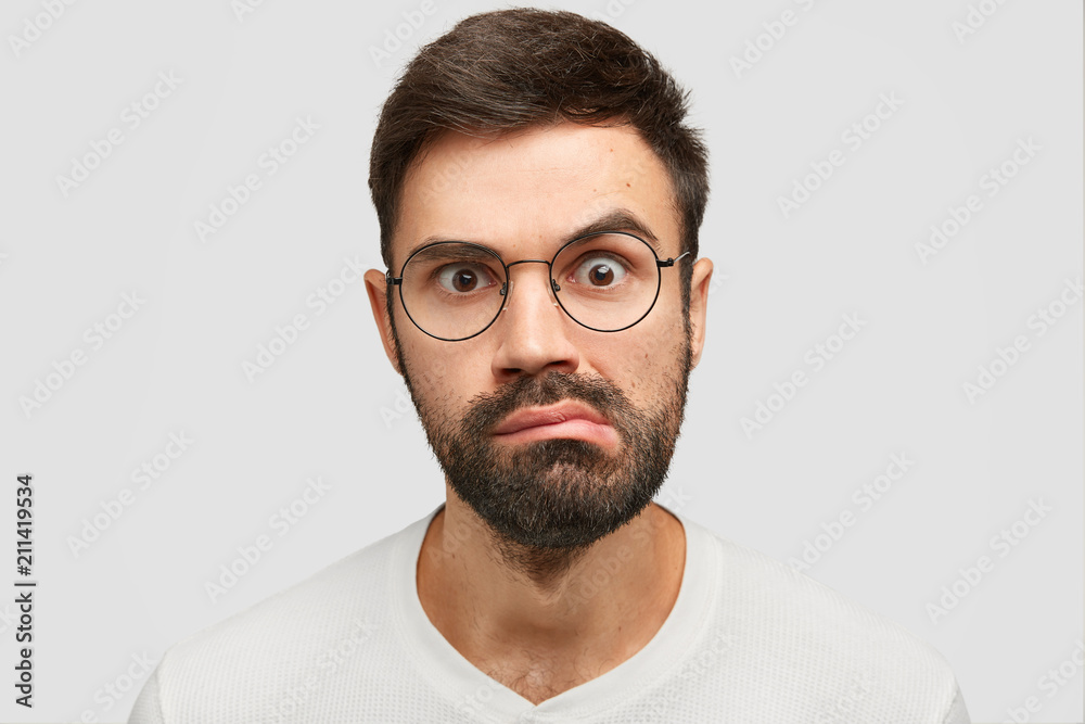 Close up portrait of attractive young male with dark stubble, looks surprisingly at camera, purses lips, stares through spectacles, has puzzled look. Shocked handsome guy has frustrated expression Close up portrait of attractive young male with dark stubble, looks surprisingly at camera, purses lips, stares through spectacles, has puzzled look. Shocked handsome guy has frustrated expression