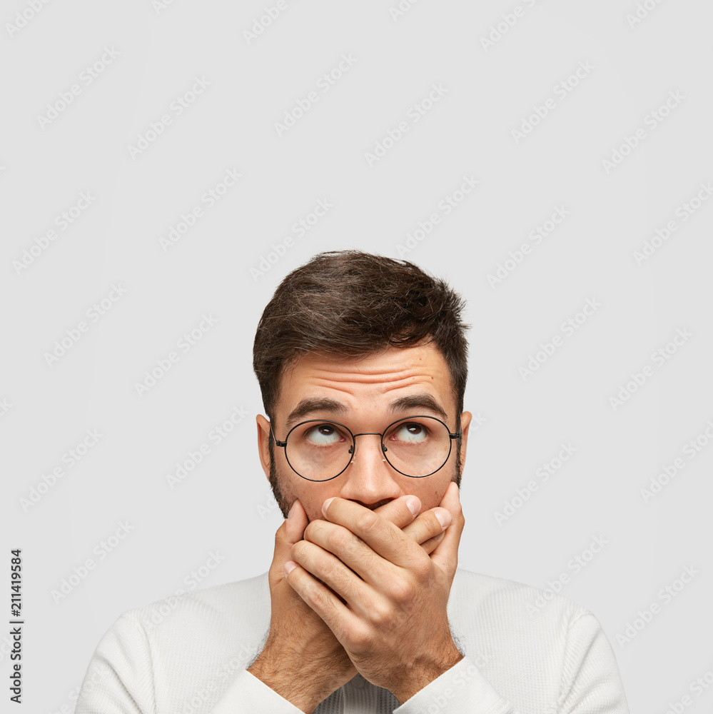 Stupefied Caucasian young male with dark hair, covers mouth with palms, looks upwards, tries to be quiet and not produce any sound, wears casual clothes. Monochrome. People and reaction concept Stupefied Caucasian young male with dark hair, covers mouth with palms, looks upwards, tries to be quiet and not produce any sound, wears casual clothes. Monochrome. People and reaction concept