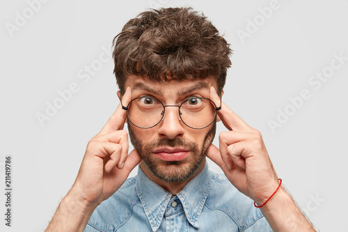 Serious bearded young Caucasian male keeps fingers on temples, tries to recollect something important, looks stressfully, focused on information, thinks over suggestion, isolated on white wall