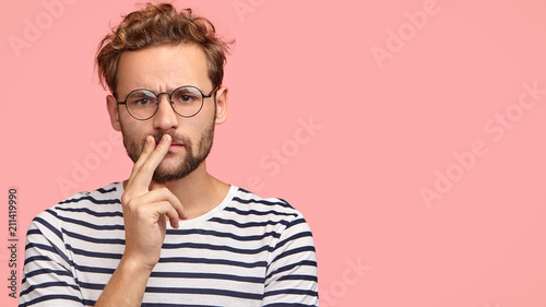 Horizontal shot of serious displeased man frowns face, keeps hand on mouth, has curly hair and stubble, wears striped t shirt, round spectacles, stands against pink background with copy space