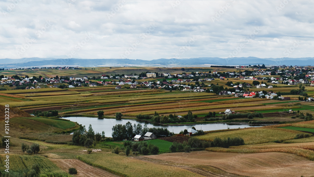 A typical natural landscape in the country of Suceava: fields, a small lake and a village of houses with red roofs, mountains on the horizon, Romania. Panoramic view.