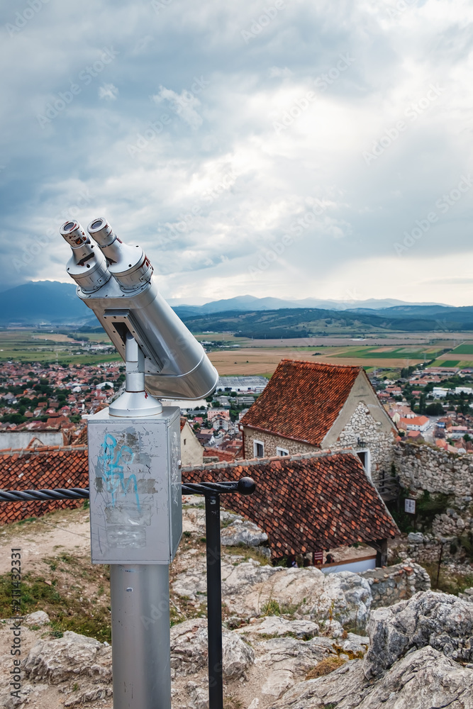 Touristic telescope look at the Rasnov medieval city from the Rasnov citadel, Brasov county, Romania. Old city with red roofs, fields and mountains under a cloudy evening sky