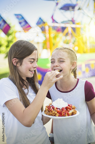 Two laughing and smiling teenage girls eating a funnel cake at an outdoor carnival or amusement park. Cute expression in this candid photo