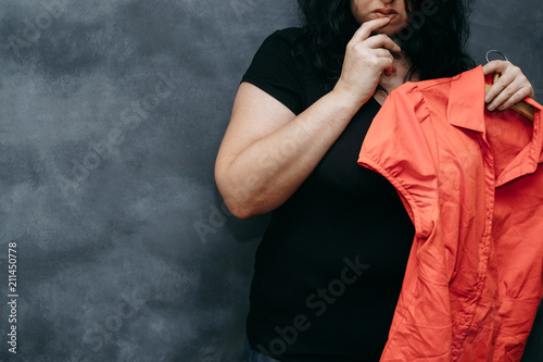 Overweight, low self-esteem, body shaming. Obese shy woman hesitating to wear vibrant colored shirt photo