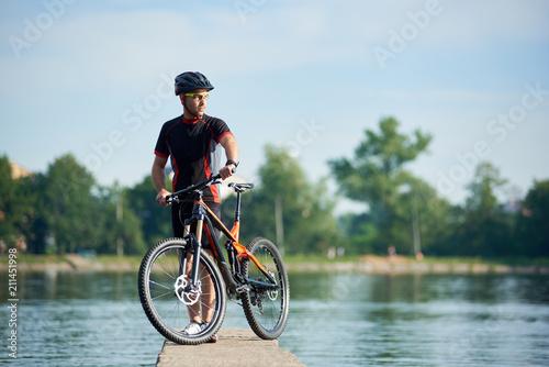 Athletic man biker in professional cycling clothing and protective helmet posing near bicycle looking far thinking about future success and achievement. Sportsman taking break at pier near lake.