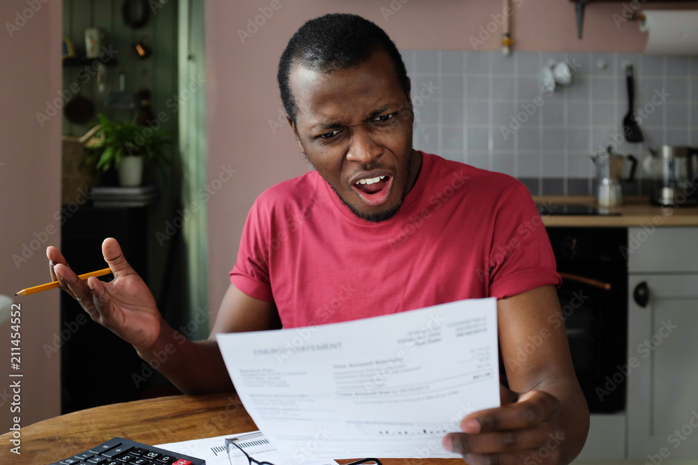 Irritated young african american man looking at utility bill, worried about troubles with mortgage payment to bank, having serious problems with debt, frustrated about high taxes Irritated young african american man looking at utility bill, worried about troubles with mortgage payment to bank, having serious problems with debt, frustrated about high taxes