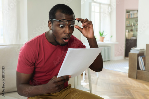 Young african american man sitting on couch in modern apartment with mouth open, holding utility bill with high rates, raised eyeglasses in wow or surprise gesture photo