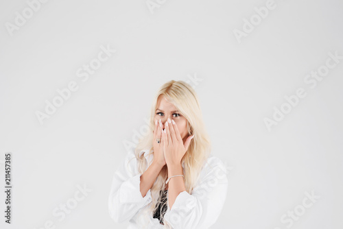 Happy blond women standing with hands near opened mouth and popping eyes. Girl with long hair in a white shirt on white isolated background in studio