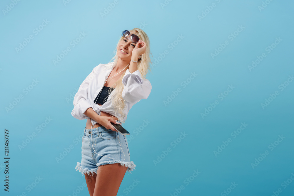 A young slender blonde girl with long hair carefree stands on a blue background and flips music on her headphones. A model in summer clothes and sunglasses smiles and imagines herself on the beach A young slender blonde girl with long hair carefree stands on a blue background and flips music on her headphones. A model in summer clothes and sunglasses smiles and imagines herself on the beach