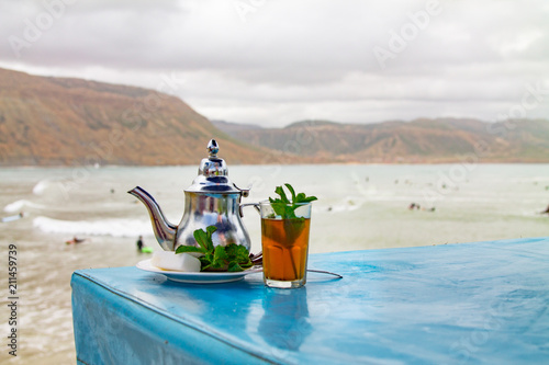 A glass of mint tea with an iron kettle, a sprig of mint and a large slice of sugar. A traditional Berber drink mint tea in the background of the Atlantic Ocean and mountains. Africa, Morocco, Agadir