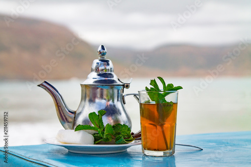 A glass of mint tea with an iron kettle, a sprig of mint and a large slice of sugar. A traditional Berber drink mint tea in the background of the Atlantic Ocean and mountains. Africa, Morocco, Agadir © Galyna Chyzh