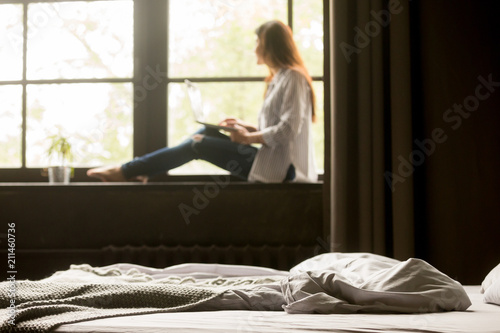 Thoughtful young woman dreaming looking in window working at laptop sitting on bedroom windowsill in cozy loft apartment, dreamy female thinking, browsing internet, relaxing at home. Focus on bed
