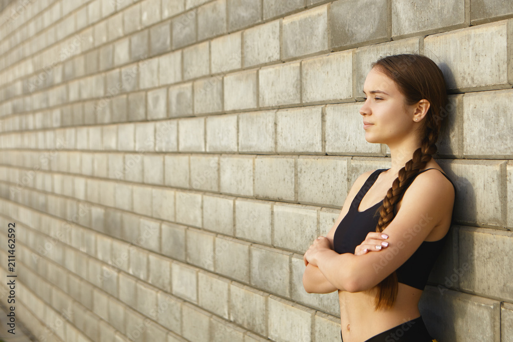 Sporty young woman relaxing after jogging workout, keeping arms folded. Picture of attractive young woman athlete resting at brick wall with copy space for your text or advertising information Sporty young woman relaxing after jogging workout, keeping arms folded. Picture of attractive young woman athlete resting at brick wall with copy space for your text or advertising information