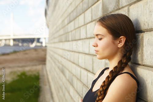 Cropped shot of stylish beautiful young sportswoman with braid leaning back agaist brick wall, having small break whie exercising outdoors, feeling tired. People, activity, health and sports concept