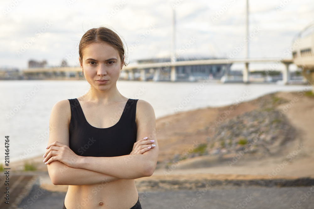Serious confident young European female runner wearing black sports top posing outdoors with sea in background, crossing arms on her chest, looking at camera. Fitness and active lifestyle concept