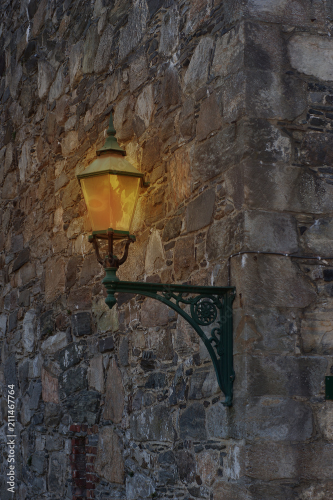 An ancient street lamp illuminating a street and a stone wall in the city An ancient street lamp illuminating a street and a stone wall in the city
