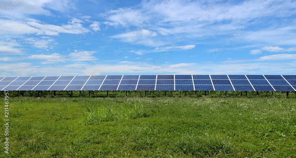 Long row of solar panels in a meadow, between green grass and clouds moving in the sky Long row of solar panels in a meadow, between green grass and clouds moving in the sky