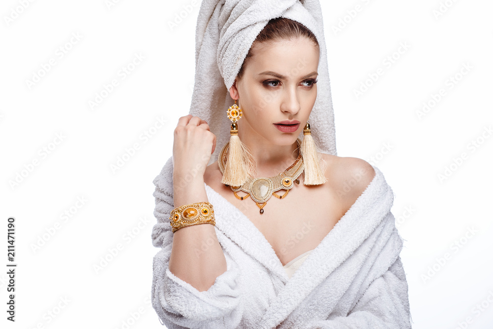 Fototapeta premium Young woman in a bathrobe and towel on her head, fashion golden jewelry, spa and care portrait, clean natural face, portrait on a background isolated