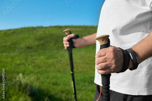 Man holding tracking sticks on green grass background. Healthy lifestyle, outdoors activities. Sportsman wearing white shirt, black trousers, handwatch. Everyday trainings on fresh air.