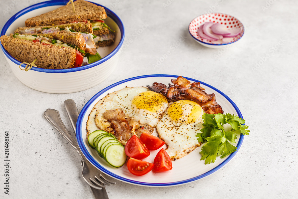 Fried eggs with bacon and a sandwich with meat, cheese and vegetables on white background. Delicious hearty breakfast.