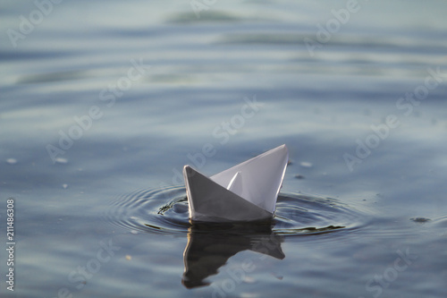 Close-up of simple small white origami paper boat floating quietly in blue clear river or sea water under bright summer sky. Freedom, dreams and fantasies concept, copyspace background.