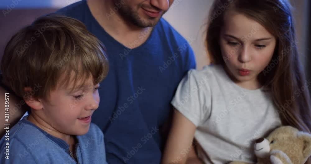 Close up of the cute kids with their father sitting in the bed in the evening and looking at the book they reading before sleeping. Indoors. Close up of the cute kids with their father sitting in the bed in the evening and looking at the book they reading before sleeping. Indoors.