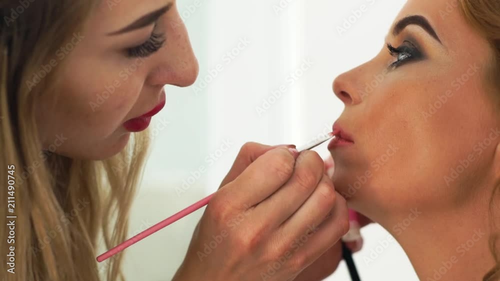 Makeup artist doing makeup lips to beautiful woman in beauty studio. Close up visagiste using brush for application lip gloss. Makeup lips on master class. Fashion and styling concept. Makeup artist doing makeup lips to beautiful woman in beauty studio. Close up visagiste using brush for application lip gloss. Makeup lips on master class. Fashion and styling concept.