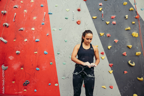 Climbing female instructor ajusting climbing equipment standing over painted in red and grey climbing wall in gym photo