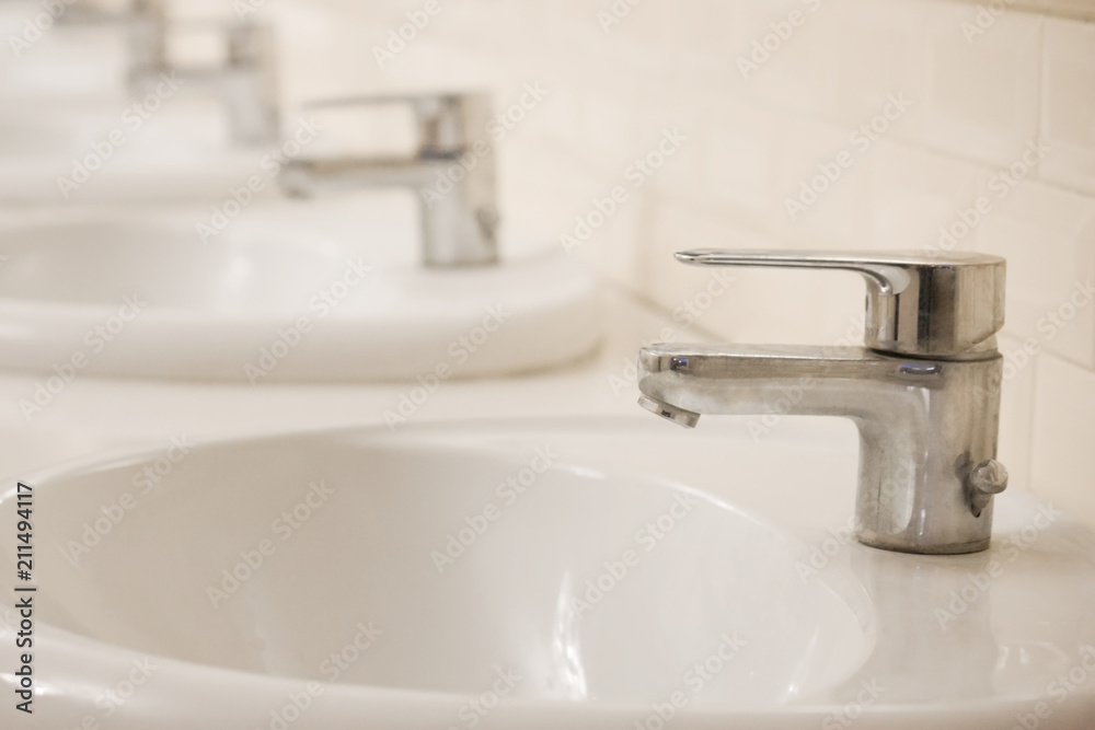 Fototapeta premium Many Closed chrome-plated faucets stand in a row in a public toilet. The room is clean and bright.